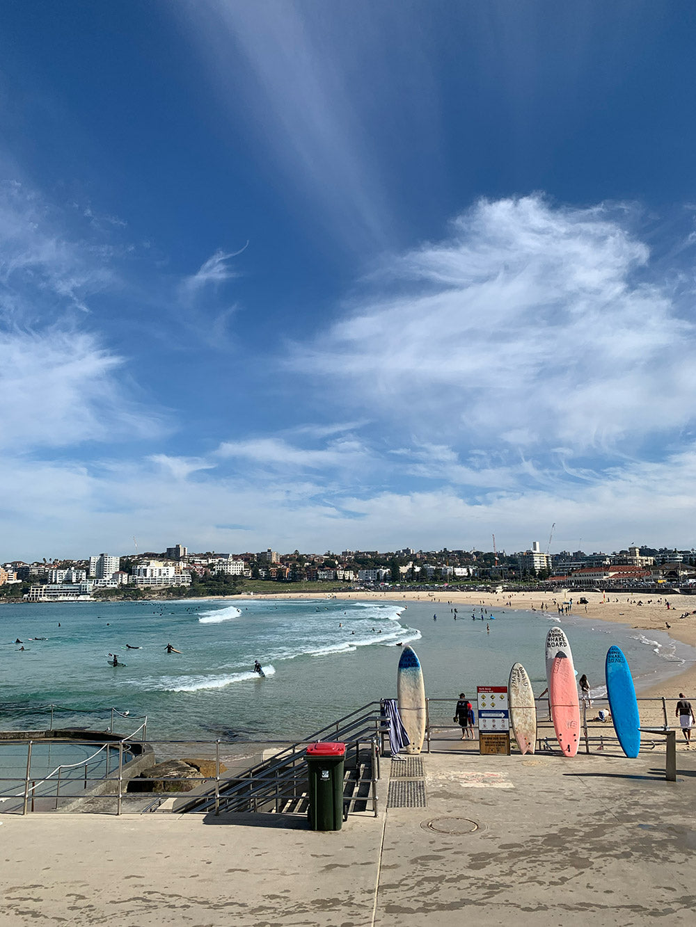 View over Bondi beach, Sydney, Australia. 4 surfboards leaning against the rail of the steps down to the beach with Bondi Bay background behind. The sky is blue with some clouds.